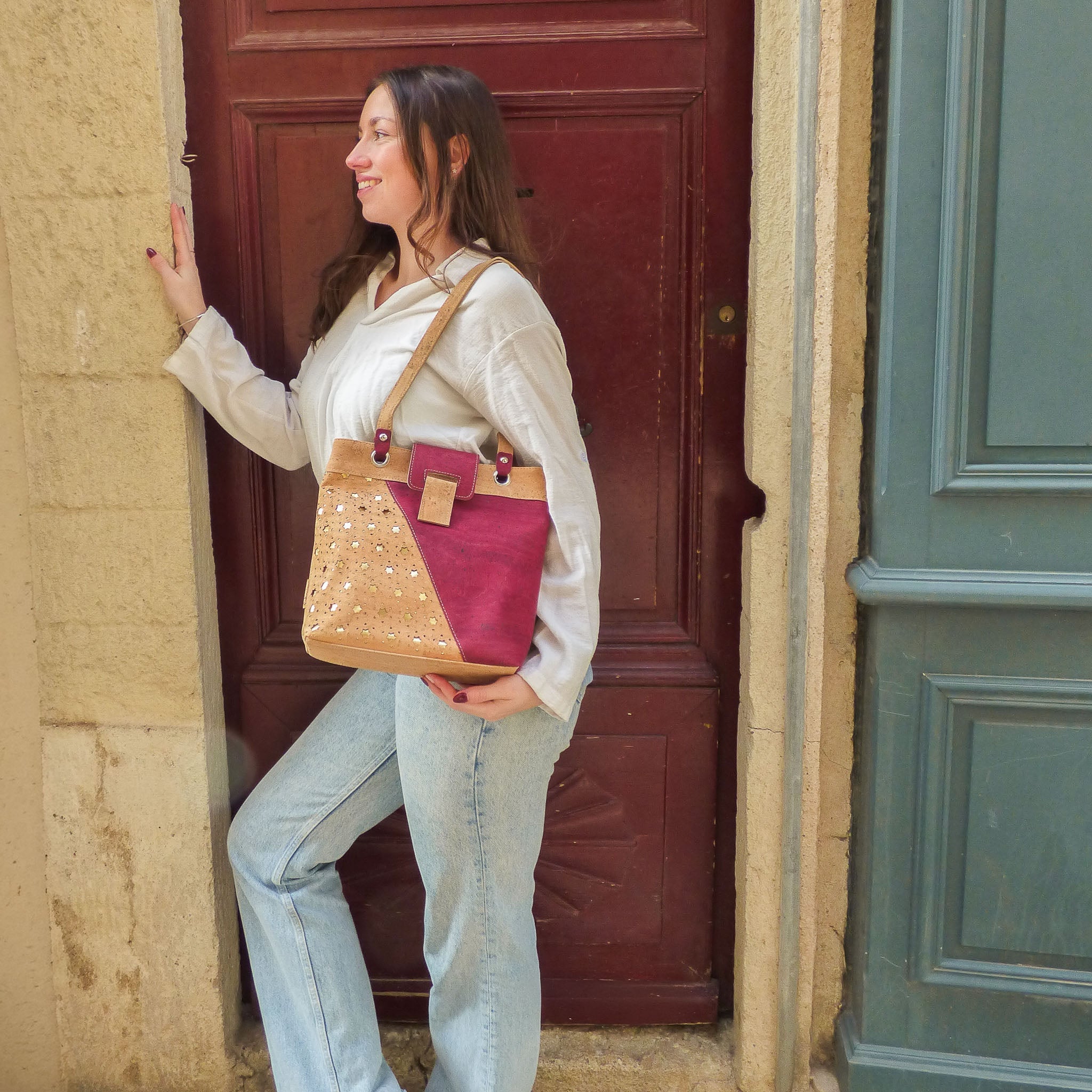Femme souriante portant un sac à main en liège bordeaux devant une porte ancienne